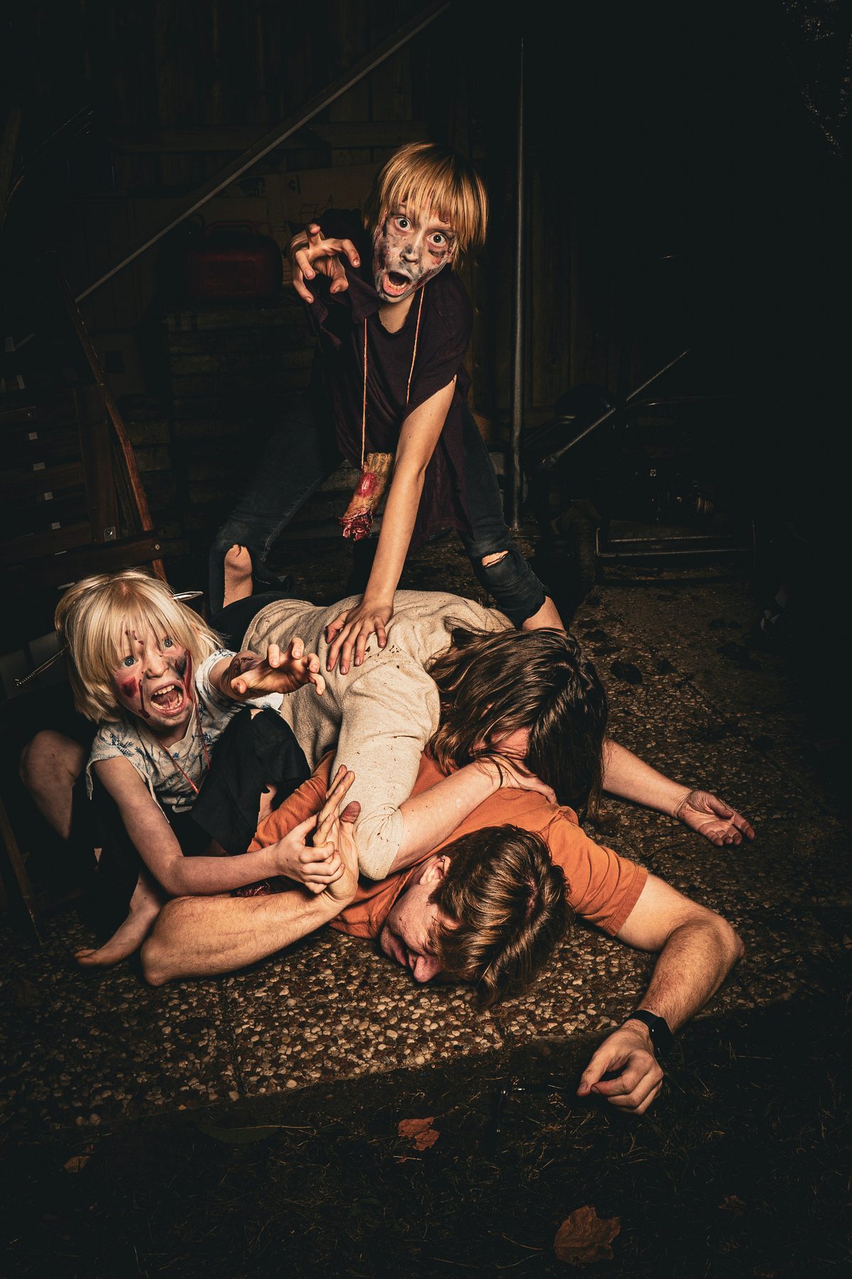 Two zombie kids in a dark shed, gleefully attacking two grown-ups sprawled face-down on the floor.