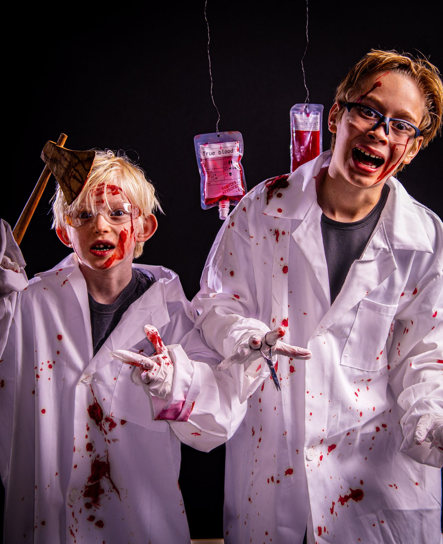 Two mad scientists in blood-spattered lab coats under hanging blood bags, the younger one with an axe in his head.